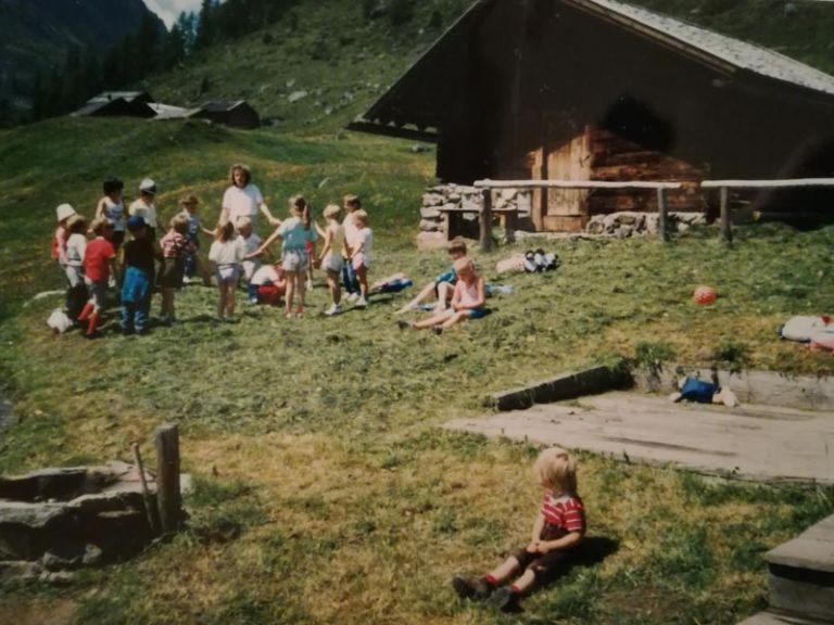 Altes Bild zeigt wie Kinder vor einer Almhütte mit einer Aufsichtsperson ein Spiel spielen